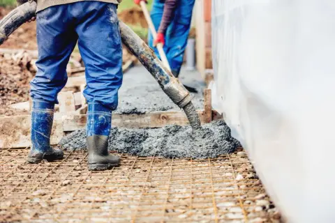 Pracownicy w niebieskich ogrodniczkach i gumowcach wylewają beton. Każdy metr sześcienny betonu to krok do solidnej budowy.