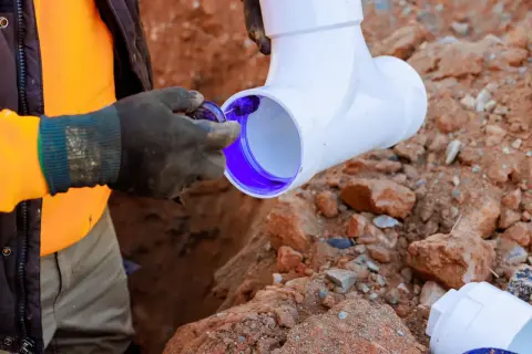 Instalator w rękawiczkach nakłada fioletowy klej na rurę PVC, przygotowując ją do montażu. Brak odpowietrzenia pionu może prowadzić do problemów z instalacją.