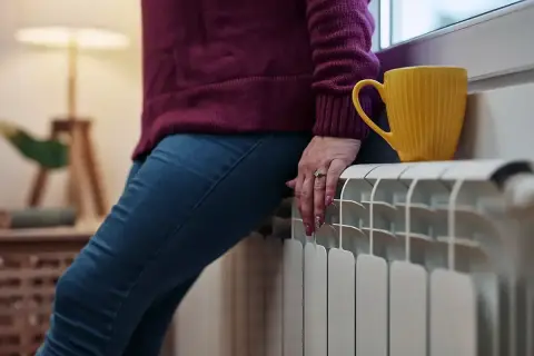 Osoba siedząca na grzejniku, obok żółty kubek. Widać, jak duża jest moc grzewcza grzejnika, ogrzewając wnętrze.